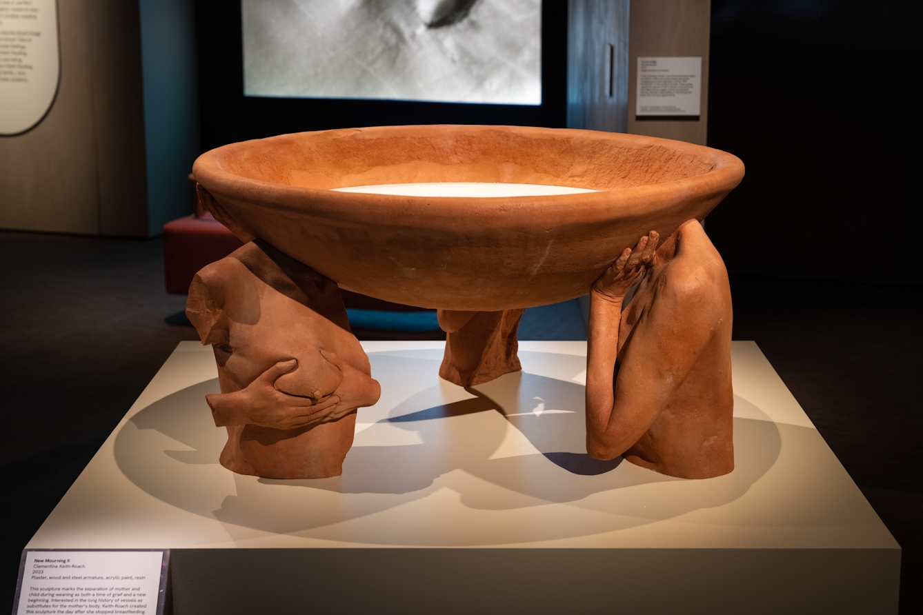Photograph of a large terracotta artwork in an exhibition space. The artwork is made up of a large bowl, supported on 3 sides by truncated terracotta torsos, one of which is holding its right breast.  Behind the artwork are framed works, video projections and caption panels.
