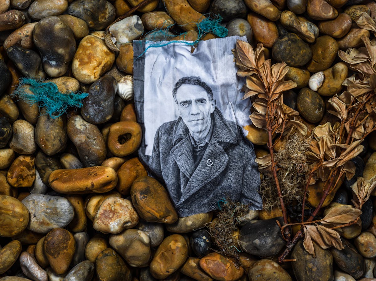 Photograph of a shingle beach made up of pebbles of all different sizes and shapes with a mix of brown and yellow hues. Nestled in the pebbles is a black and white photographic print, soaked in sea water such that it has taken on the shape of the stones beneath. Staring out from the print is the gentle face of Derek Jarman, looking intensely to camera. He wears a think woollen coat with a small silver heart broach pinned to the lapel. Surrounding the print on the stones are sea shells, sprigs of beachfront vegetation and the frayed blue nylon threads of fishing nets.