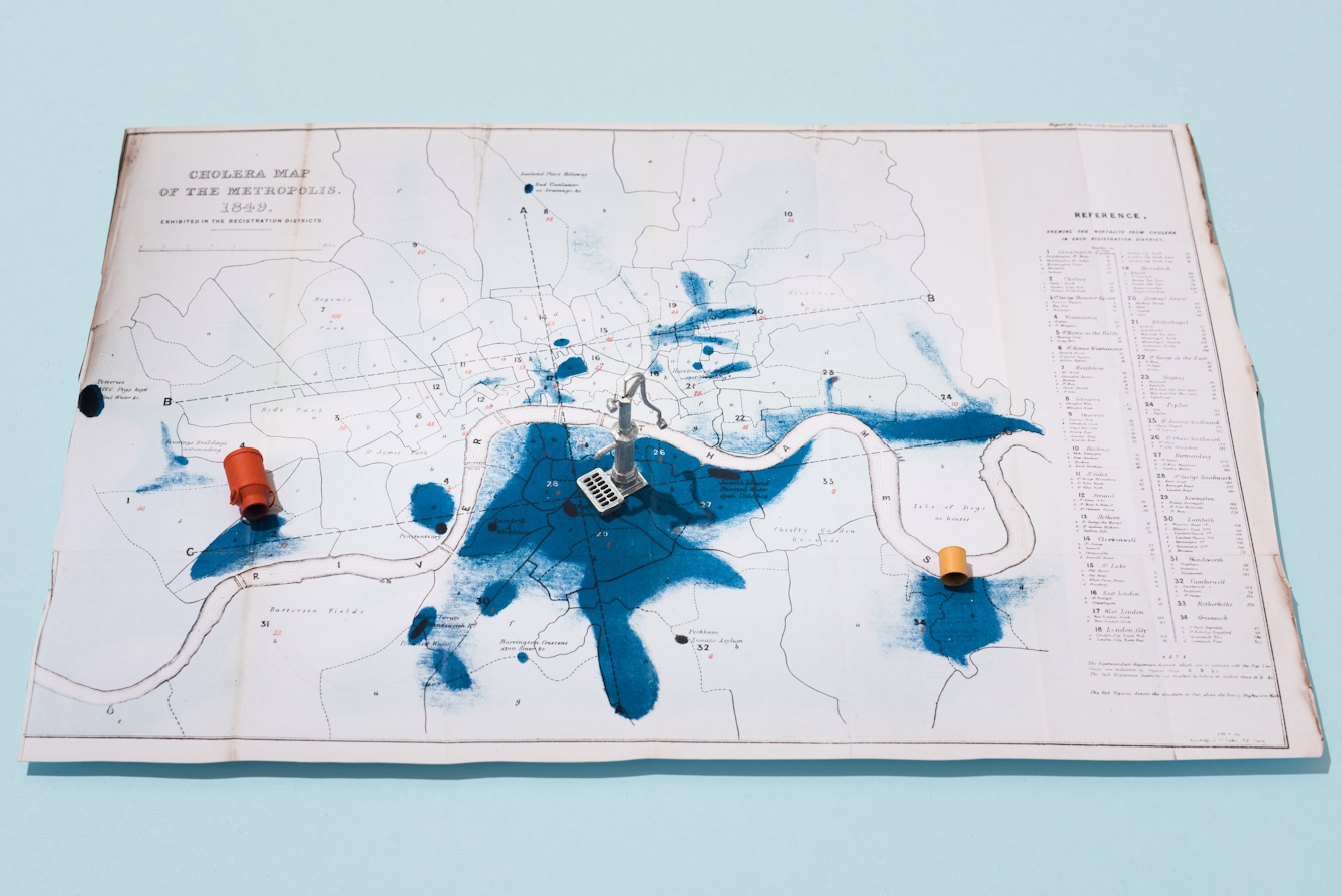 Photograph of a 'heat' map of London dated 1849, showing areas of cholera outbreaks in the city. The map is photographed at an angle, receding away from the camera. The map is slightly raised off the light blue background, casting a small shadow. A three-dimensional model water pump as well as buckets of water appear across the map's surface to illustrate the cause as being dirty water.