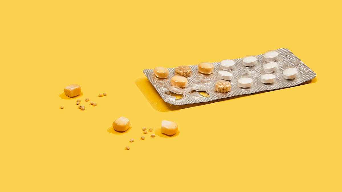 Photograph of a foil medicine pill blister pack sitting on a bright yellow background. The blisters on the right side of the pack contain white tabloid pills. The blisters on the left of the pack contain a single kernel of corn or a collection of small grains of maize. A couple of the blisters are empty and there are 3 kernels of corn and a scattering of maize grains spread out on the yellow background.