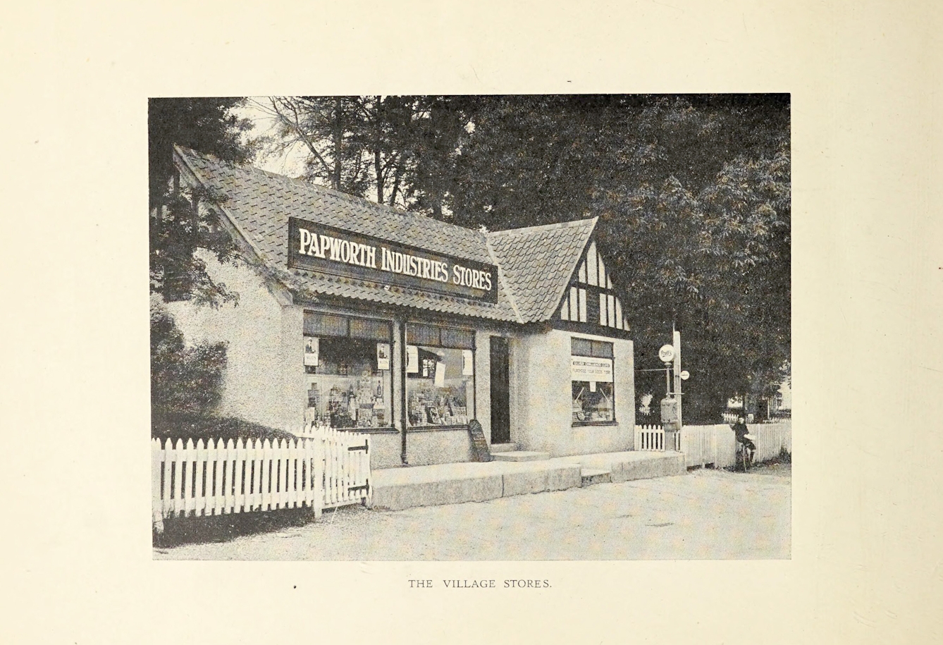 Black and white phtograph of the Papworth Industries Stores, a small village store with white picket fence on either side of it and large trees behind it. A boy on a bicycle leans against the fence to the right of the store.