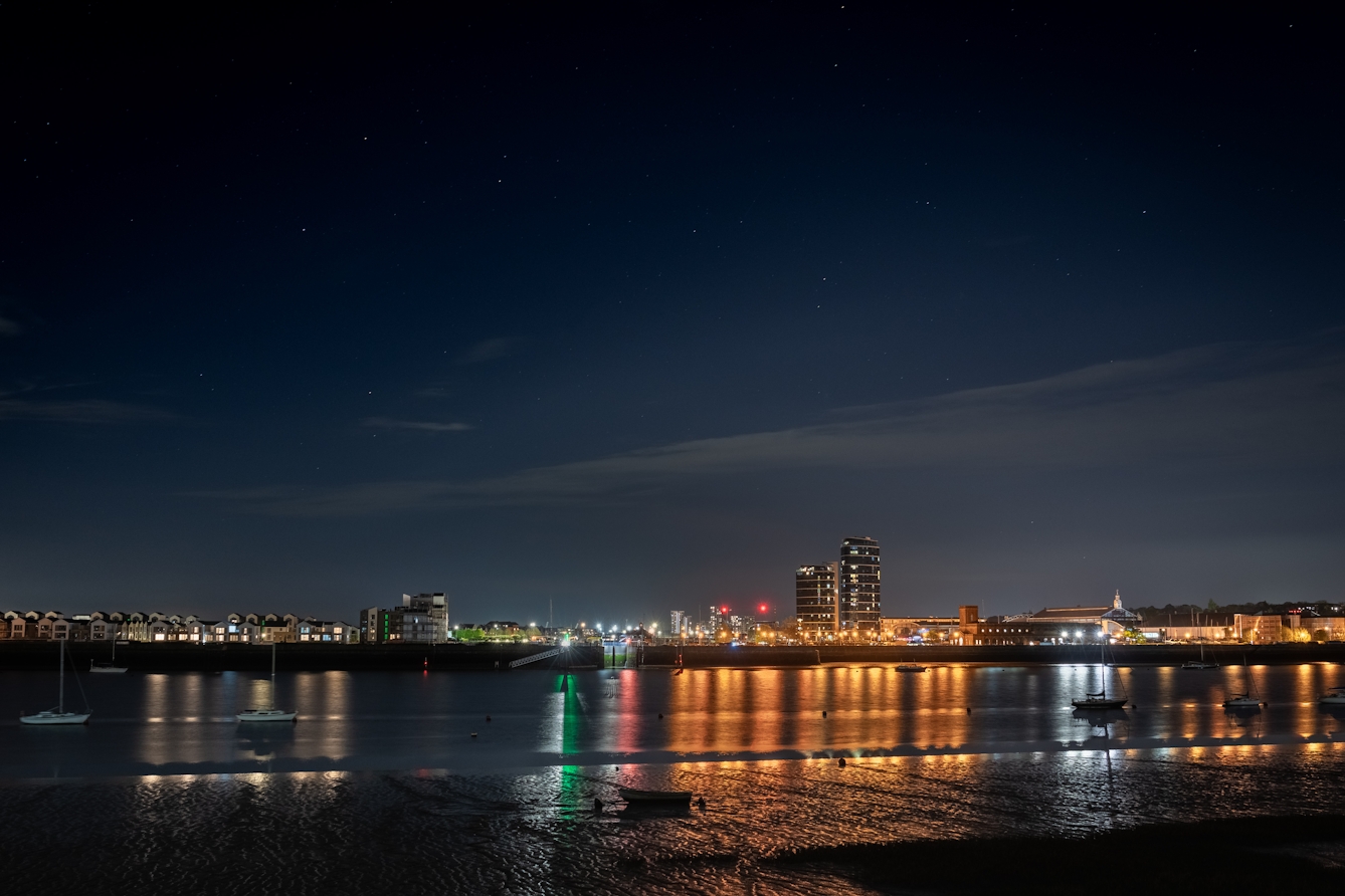 A photograph of an urban development at night and shot from the other side of a river. The development is illuminated by a street lights and from the buildings themselves, both of which are reflected in the water. The sky fades from near complete darkness, with stars visible, to a misty blue as it gets nearer to the buildings.