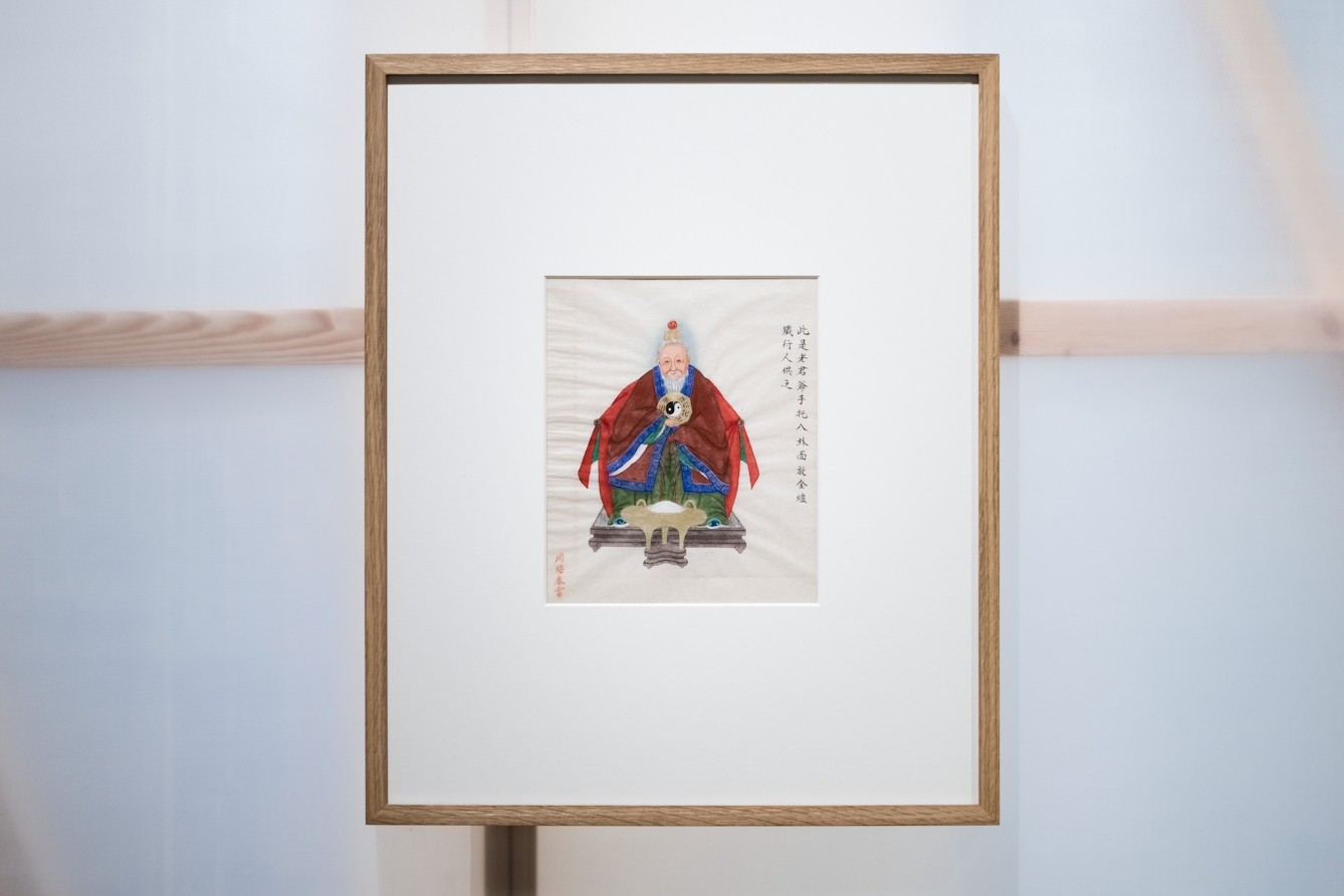 Photograph in an exhibition gallery showing a section of a translucent wall revealing the wooden structure behind, on which a framed print in a wooden frame has been hung. In a window mount within the frame is a colourful drawing of Kau Jun, the 'venerable gentleman', wearing traditional costume, holding the 'Eight Trigrams' and 'Yin and Yang' symbol.