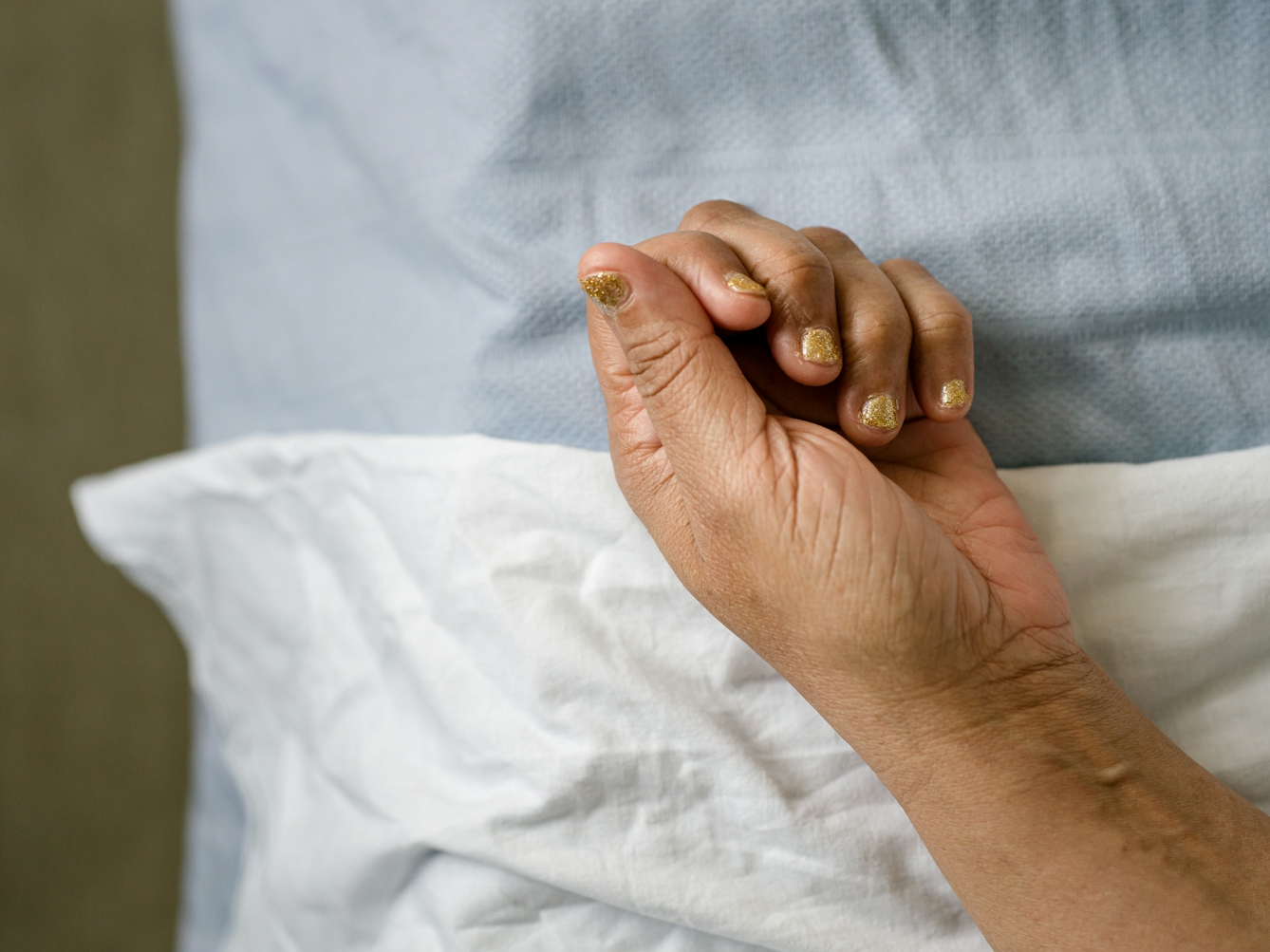 Photograph of the hand of a patient with gold glitter painted fingernails, resting on white and light blue hospital bedsheets.