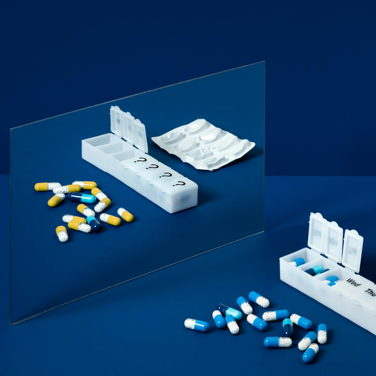 A photograph of a mirror on a blue background. In the foreground is a pill organiser, labelled with the days of the week, and loose blue and white pills. In the reflection of the mirror the pills are yellow.