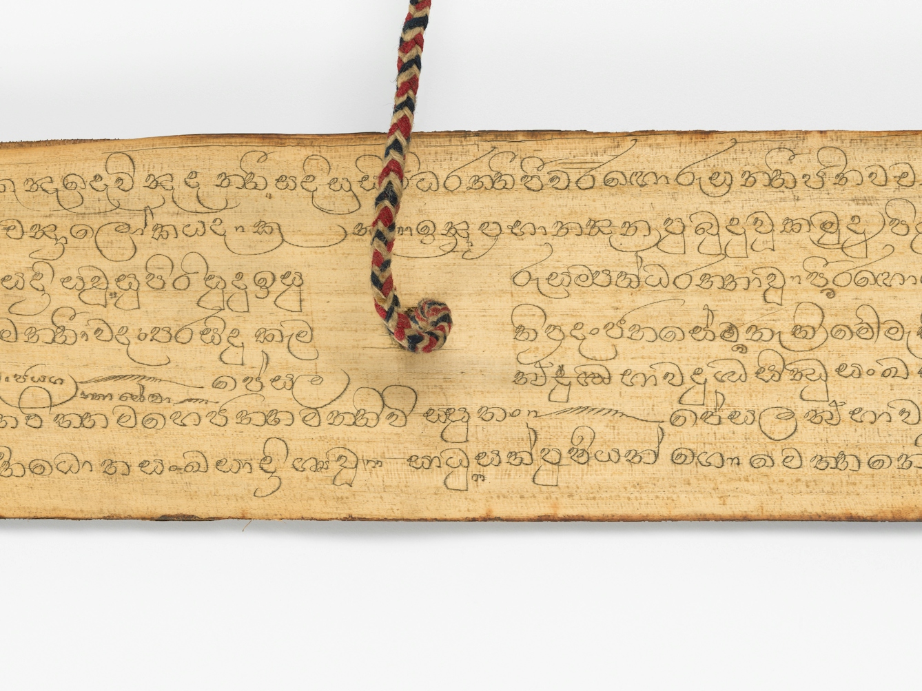 Detail of a page from a palm leaf manuscript with inscribed text in Sinhala. A red, whilte and blue string emerges from a hole in the centre of the leaf, holding the manuscript together.