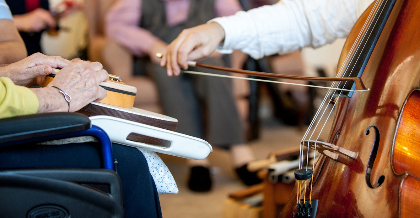 A photograph focusing in on two people sitting opposite each other, showing only their hands. The elderly woman on the left is holding some instruments on her lap, while the person opposite is holding a bow and playing the cello.