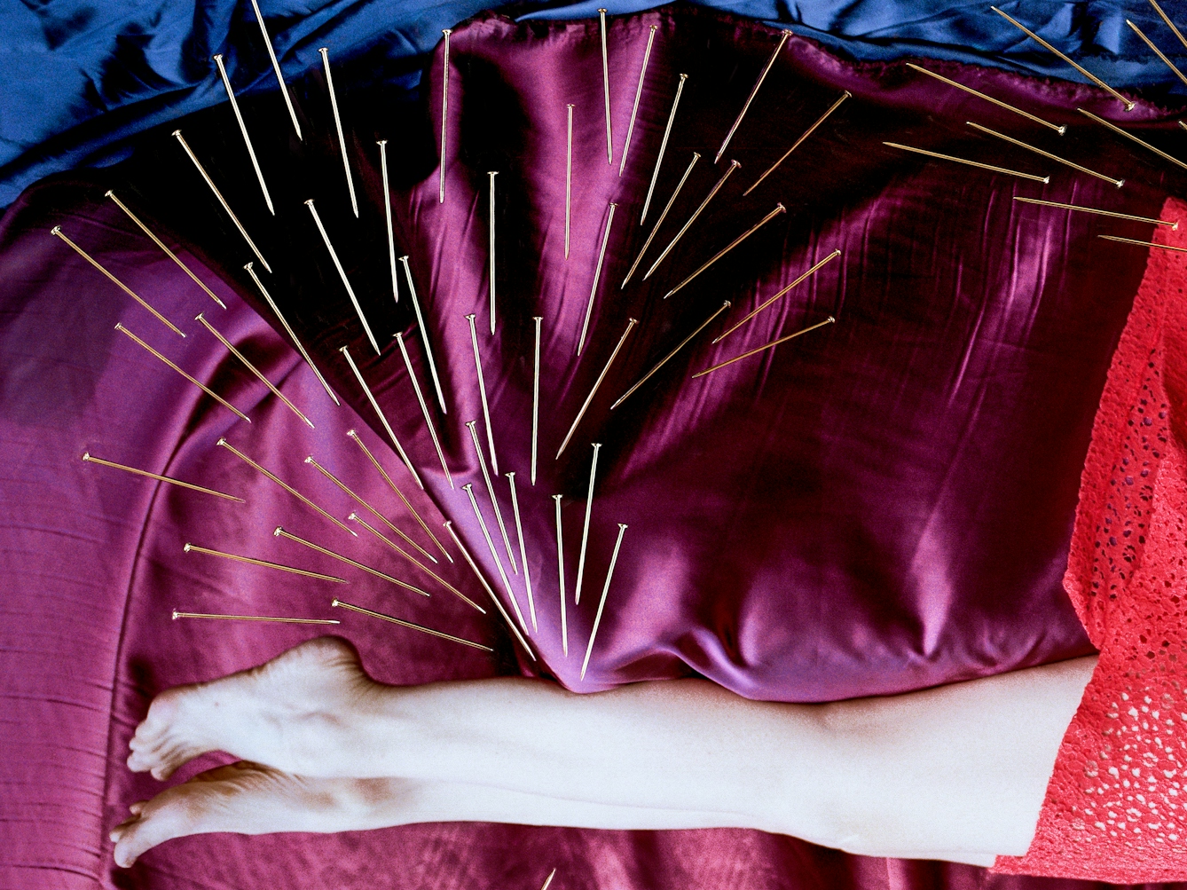 Detail from a larger artwork created with a colour photographic print of a female figure in a bright red dress, set against a purple and blue draped silk background. She is lying horizontally in the frame, face down, caught as if in mid fall. Her body is surrounded by groups of dress pins, laid on top of the photographic print. The pins are arranged as if they are a flight of arrows directed at her body. One group attacks her legs from above.