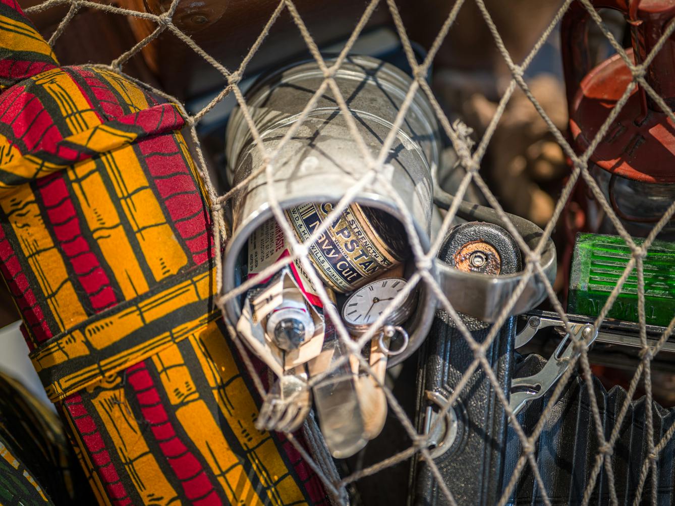 Photograph of a close-up detail of the net being carried by a life-size artwork of a figure resembling an astronaut. Within the net is a collection of objects, including a pocket watch, a camera, a knife and fork and a tin of tobacco.