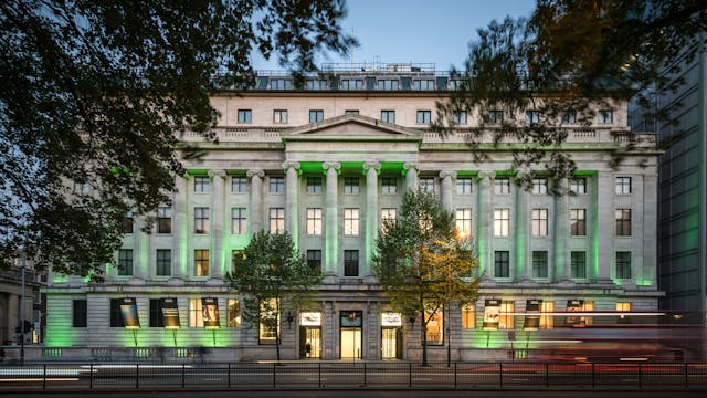 Photograph of the front elevation of the Wellcome Collection building at dusk. The building is illuminated with a series of up-lighters, flooding the facade with green light. In the foreground at ground level are blurred streaks of red and yellow from the headlights and tail lights of passing vehicles. The leaves of trees to the left and right of the image frame the building and are themselves blurred as a result of the wind and long photographic exposure.