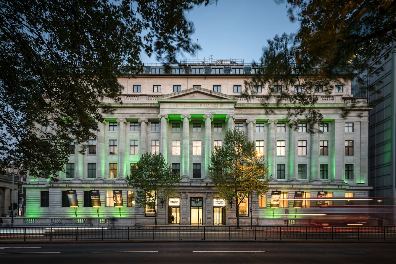 Photograph of the front elevation of the Wellcome Collection building at dusk. The building is illuminated with a series of up-lighters, flooding the facade with green light. In the foreground at ground level are blurred streaks of red and yellow from the headlights and tail lights of passing vehicles. The leaves of trees to the left and right of the image frame the building and are themselves blurred as a result of the wind and long photographic exposure.