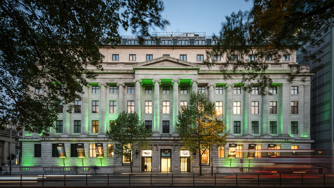 Front view of the Wellcome Collection building on Euston Road, lit green at dusk and framed by trees.