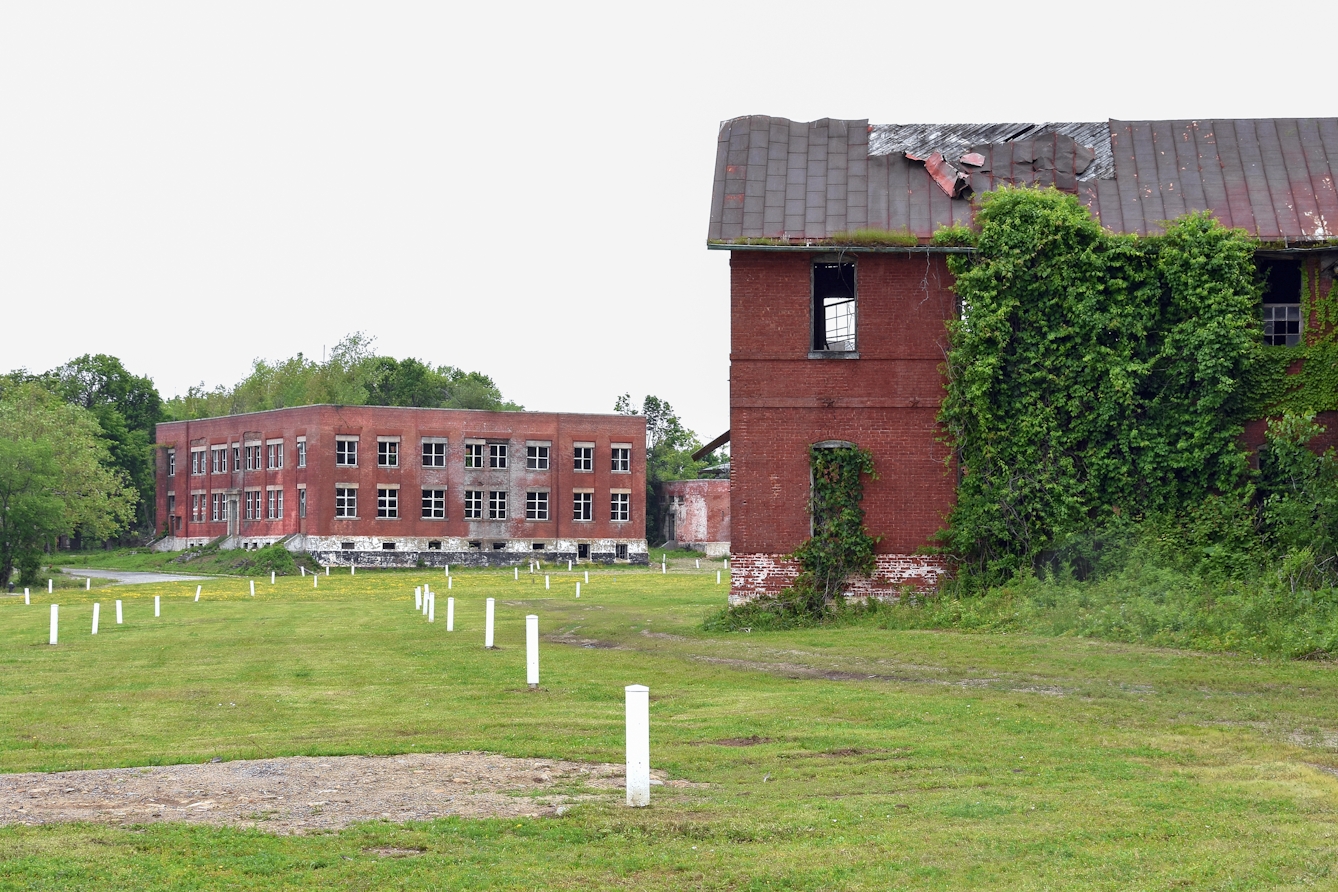 Photograph of two red brick buildings in a state of disrepair, with broken windows and disintegrating roofing materials. On the cut grass next to them are waist high white wooden rectangular posts spread in rough lines across the ground.