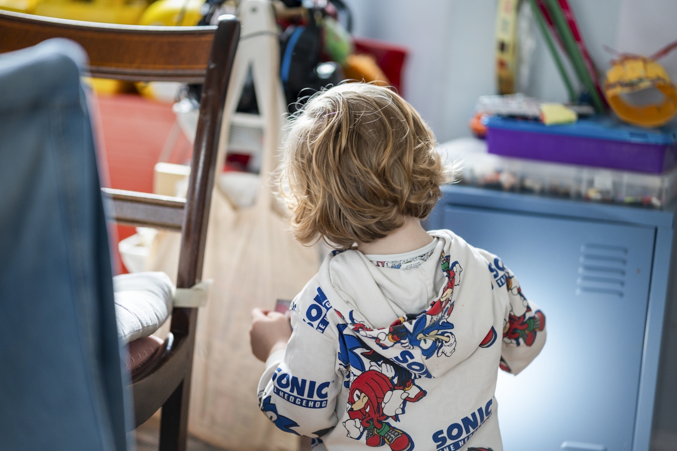 A photograph of a child, the child is facing away surrounded by toys and wearing a hoody with cartoon characters graphics.