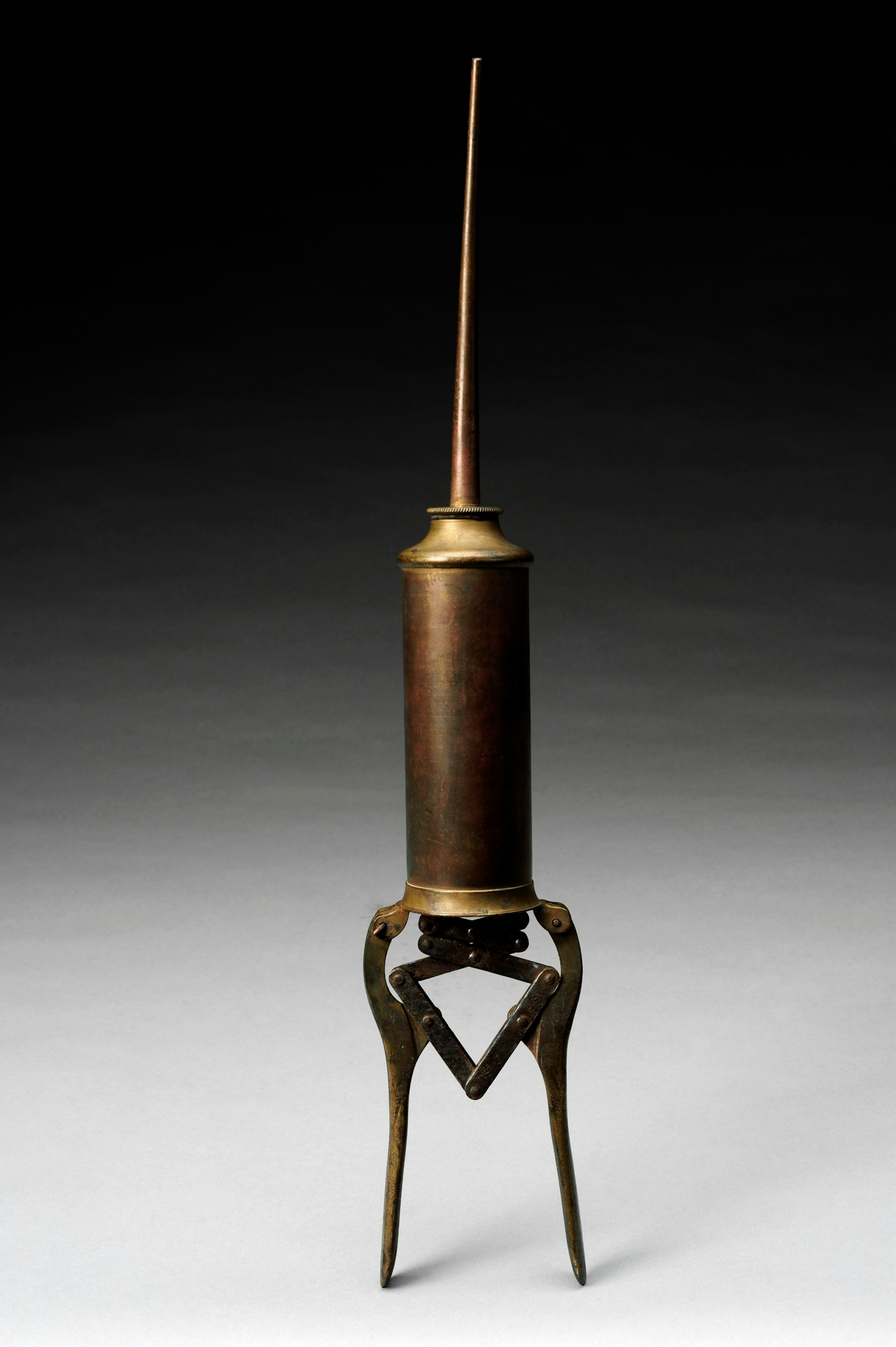 Photograph of a brass enema syringe standing vertically on a background that graduates from white at the bottom of the frame to black at the top.