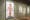 Photograph of a galley wall made of green stained wood, at an angle. On the wall are 3 artworks receding into the distance. The artwork closest to the camera shows a full length photograph of a naked woman which has been illustrated over the surface with colourful drawings.
