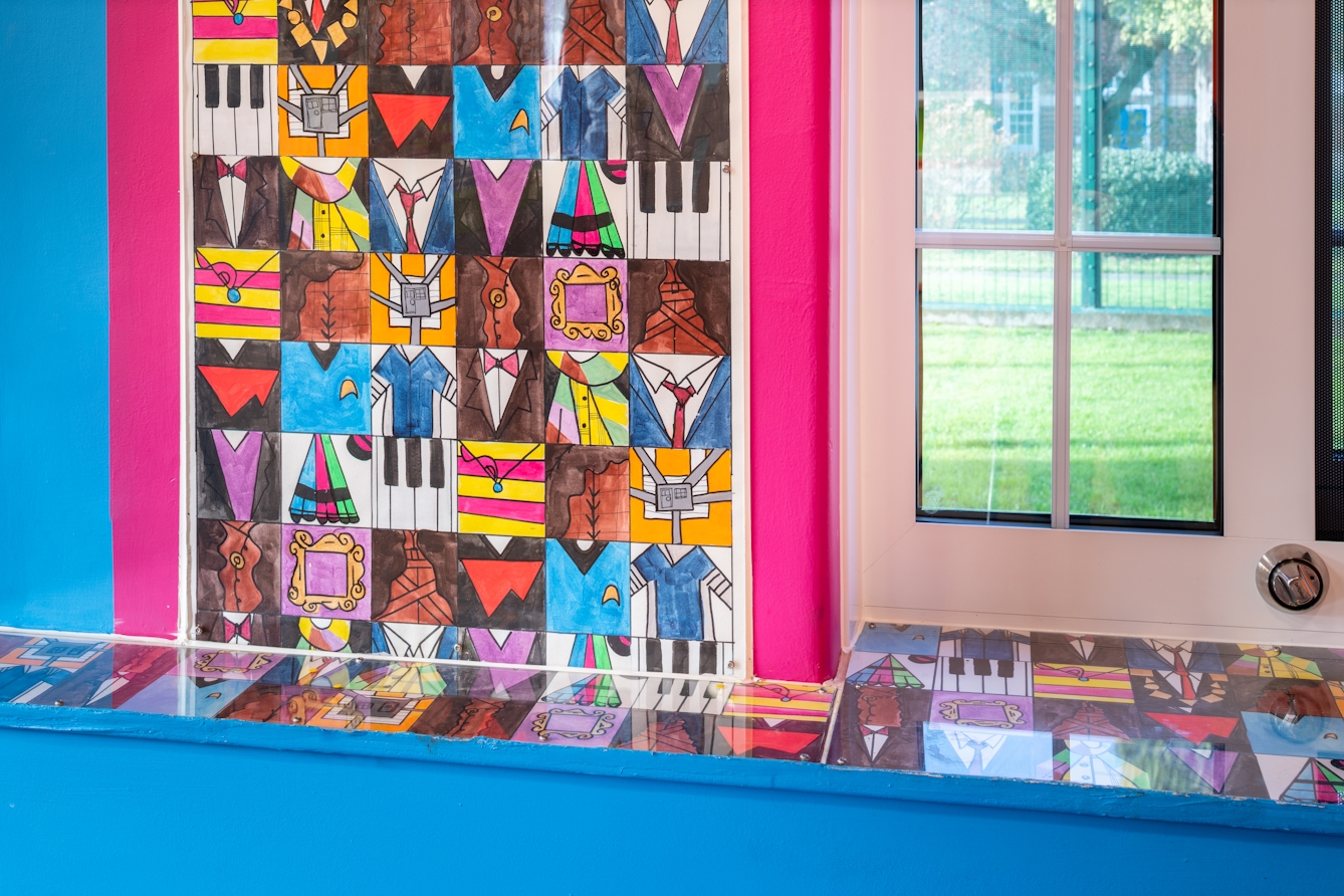 A close up photograph showing the corner of a large colourful mural on a bright pink coloured section of wall. The mural is comprised of small squares featuring simple colourful graphics including piano keys, clothed torsos and picture frames. Similar images run along the top of a narrow cupboard painted blue below. A white PVC window is visible on the right-hand side of the image.