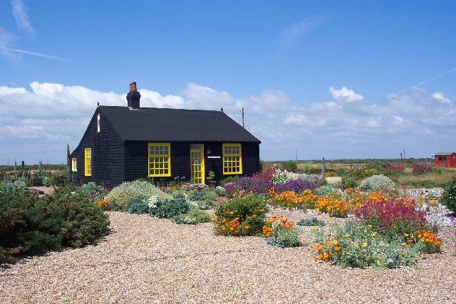 Colour photograph of a single-storey house surrounded by a garden. The house is black with bright yellow window frames and a yellow door. The garden is gravel, with colourful plants, flowers and bushes.