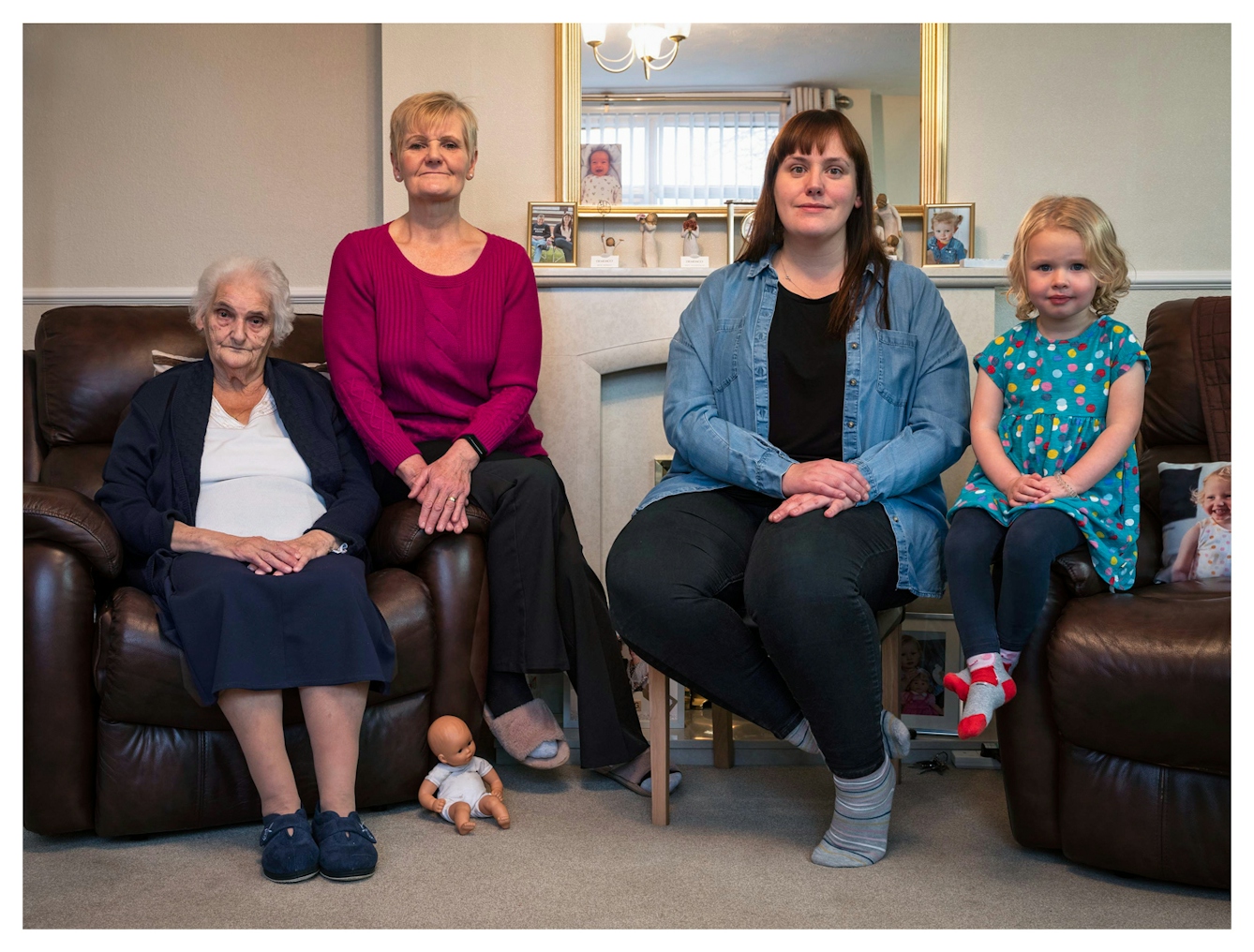 Family group photograph showing four generations of the same family. The great-grandmother sits on the left in a large armchair, by her side sits the grandmother. Next to her sits the granddaughter and next to her the great-granddaughter who is perching on the arm of an armchair. They are all seated on chairs in a living room and each of them has their hands crossed in their laps in the same way. They all look straight into the camera.