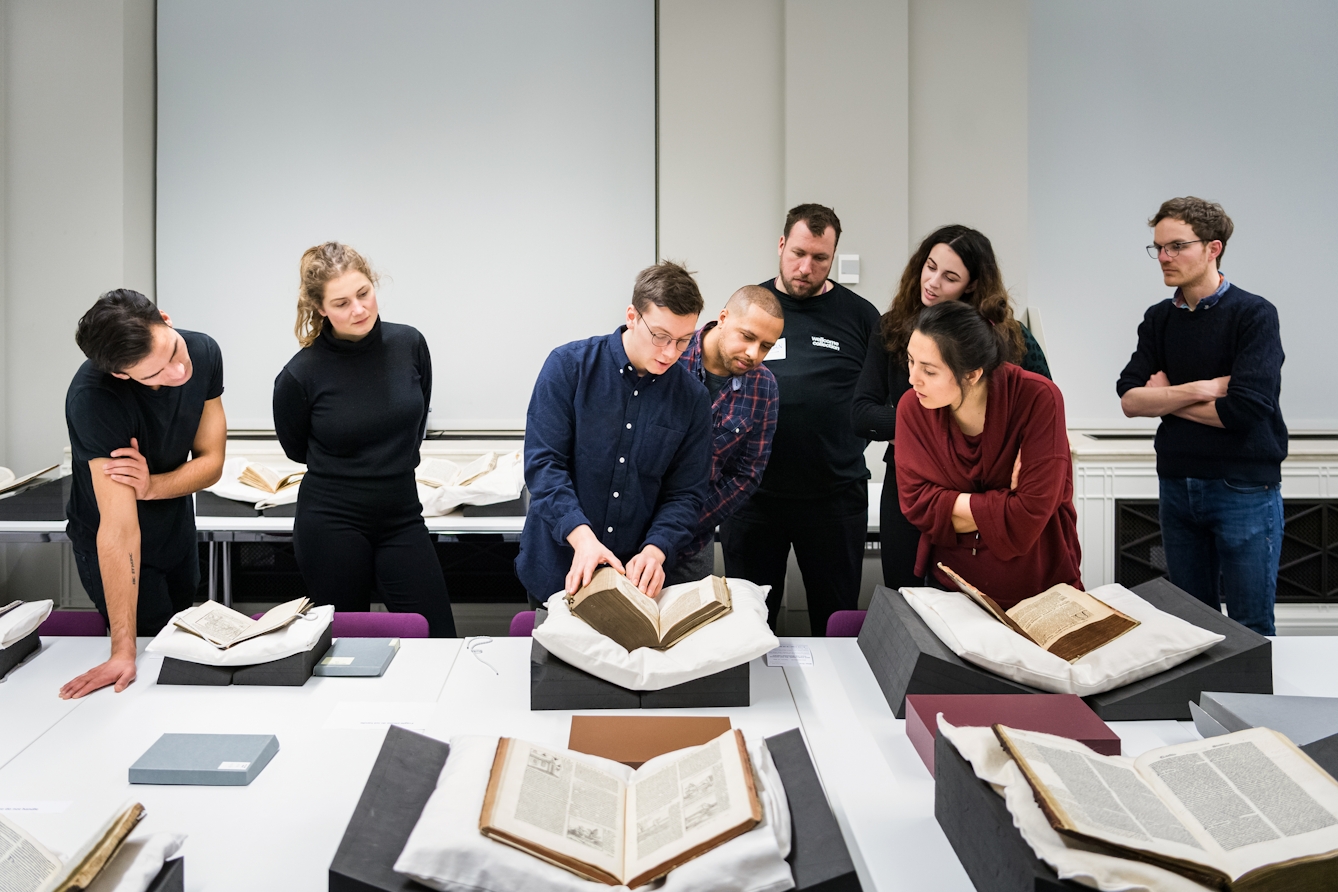 Photograph of a group of people gathered around a large table which is laid out with several old and fragile manuscripts resting on book supports. The person in the centre of the frame is touching one of the manuscripts and appears to be explaining something to the surrounding group.