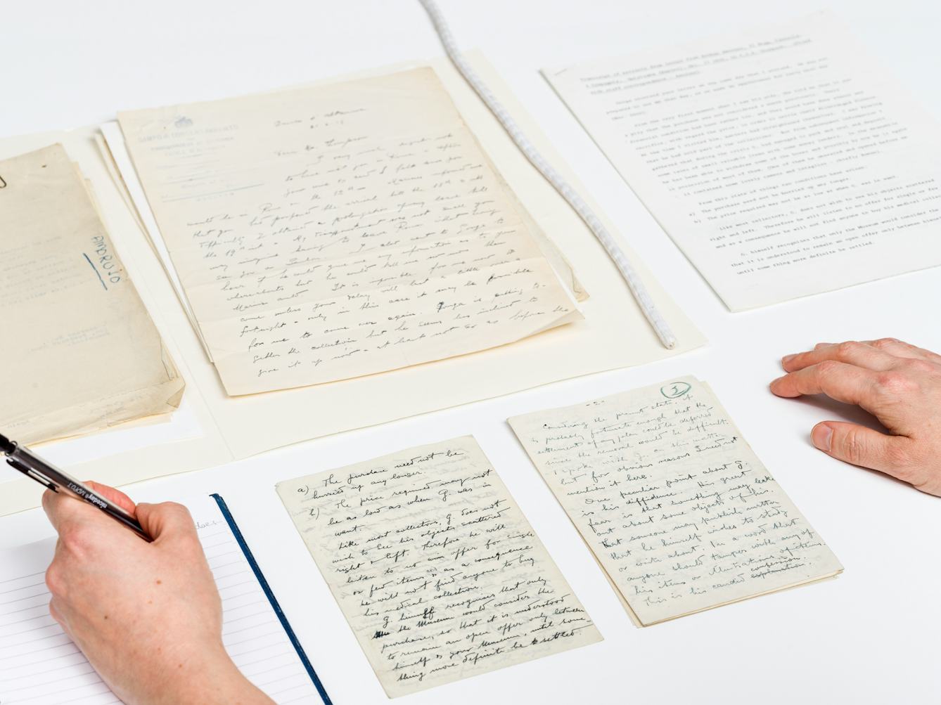 Photograph of several sheets of hand written letters and a typed letter laid out on a white table top in a careful and considered manner. A pair of hands are visible in the bottom left and right corners of the image, the right hand resting on the table, the left hand holding a pencil, writing in a lined notepad.