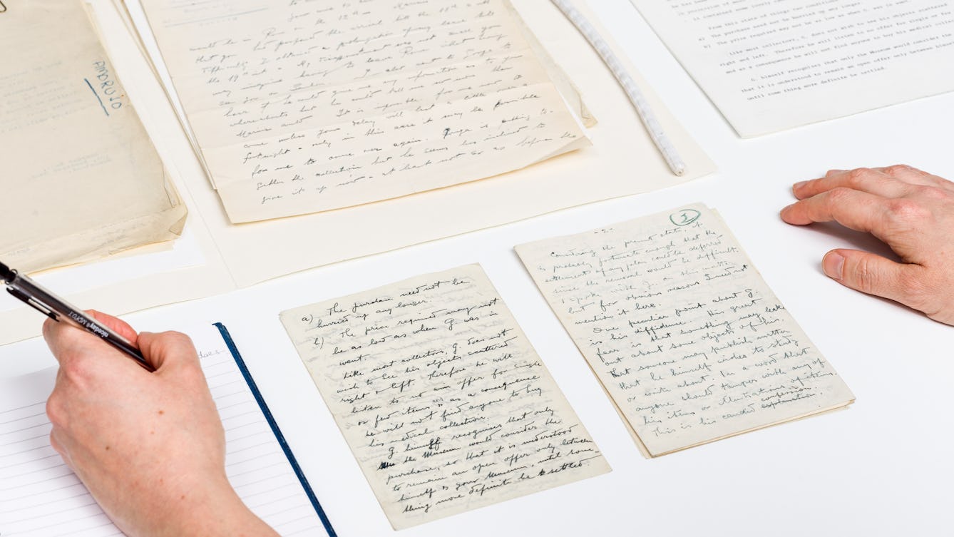 Photograph of several sheets of hand written letters and a typed letter laid out on a white table top in a careful and considered manner. A pair of hands are visible in the bottom left and right corners of the image, the right hand resting on the table, the left hand holding a pencil, writing in a lined notepad.