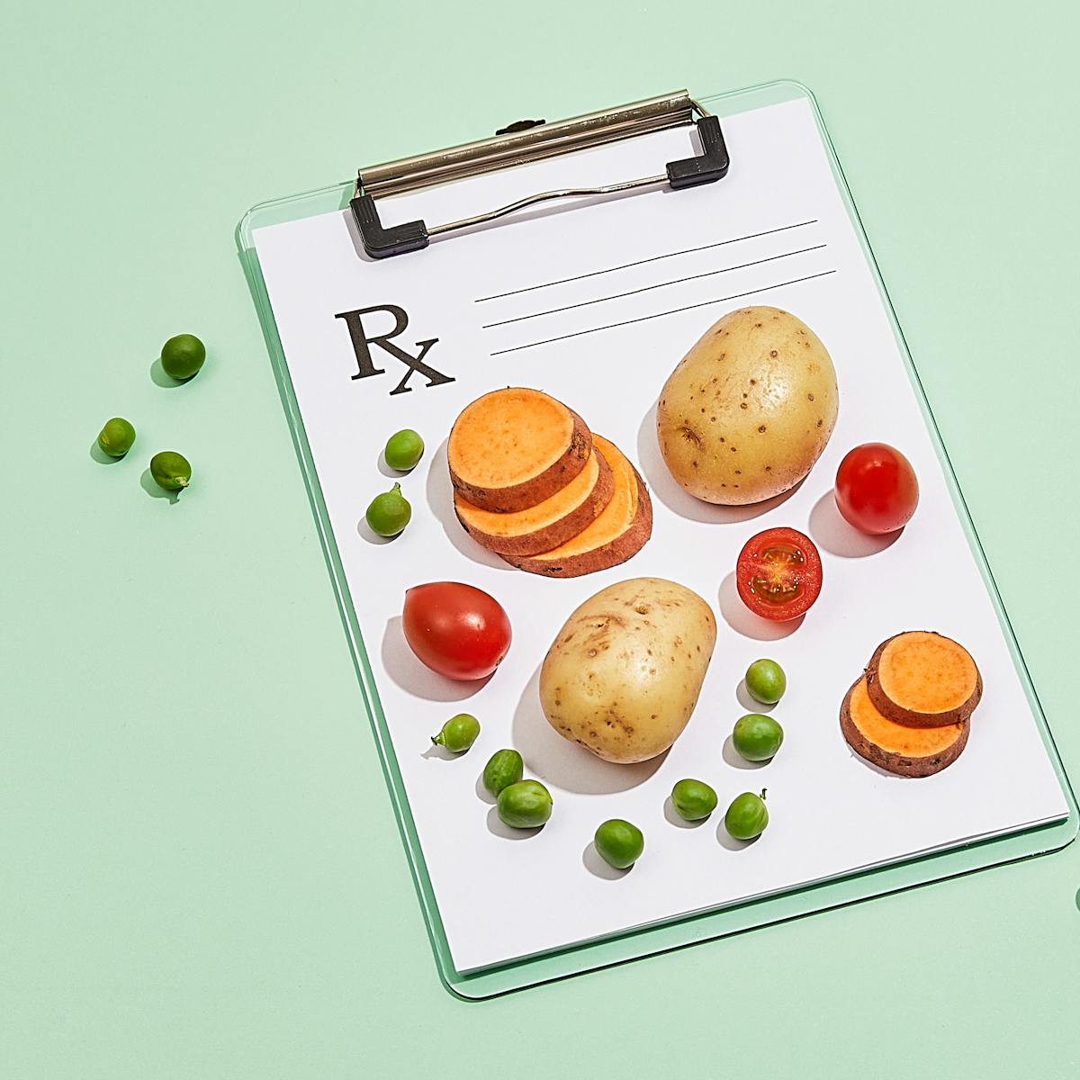 Photograph of a clipboard with a blank doctor's prescription note attached to it, resting on a light green background. On top of the paper prescription are two whole potatoes, two stacks of sliced sweet potato, whole and halved cherry tomatoes and green peas. Loose on the background, to the left and right of the clipboard are a scattering of loose green peas.