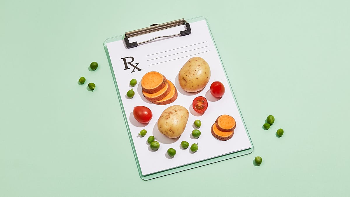 Photograph of a clipboard with a blank doctor's prescription note attached to it, resting on a light green background. On top of the paper prescription are two whole potatoes, two stacks of sliced sweet potato, whole and halved cherry tomatoes and green peas. Loose on the background, to the left and right of the clipboard are a scattering of loose green peas.