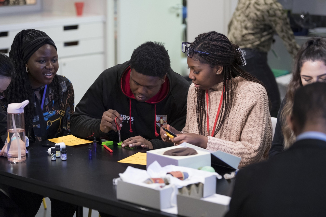 Photograph of young people sitting at a table. On the table are paper, pens and other craft materials. The young people are collaborating to make something out of these materials. 