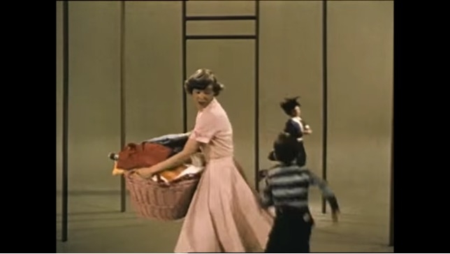 Still image from film featuring a woman in a pink dress carrying a basket of laundry and two children near her