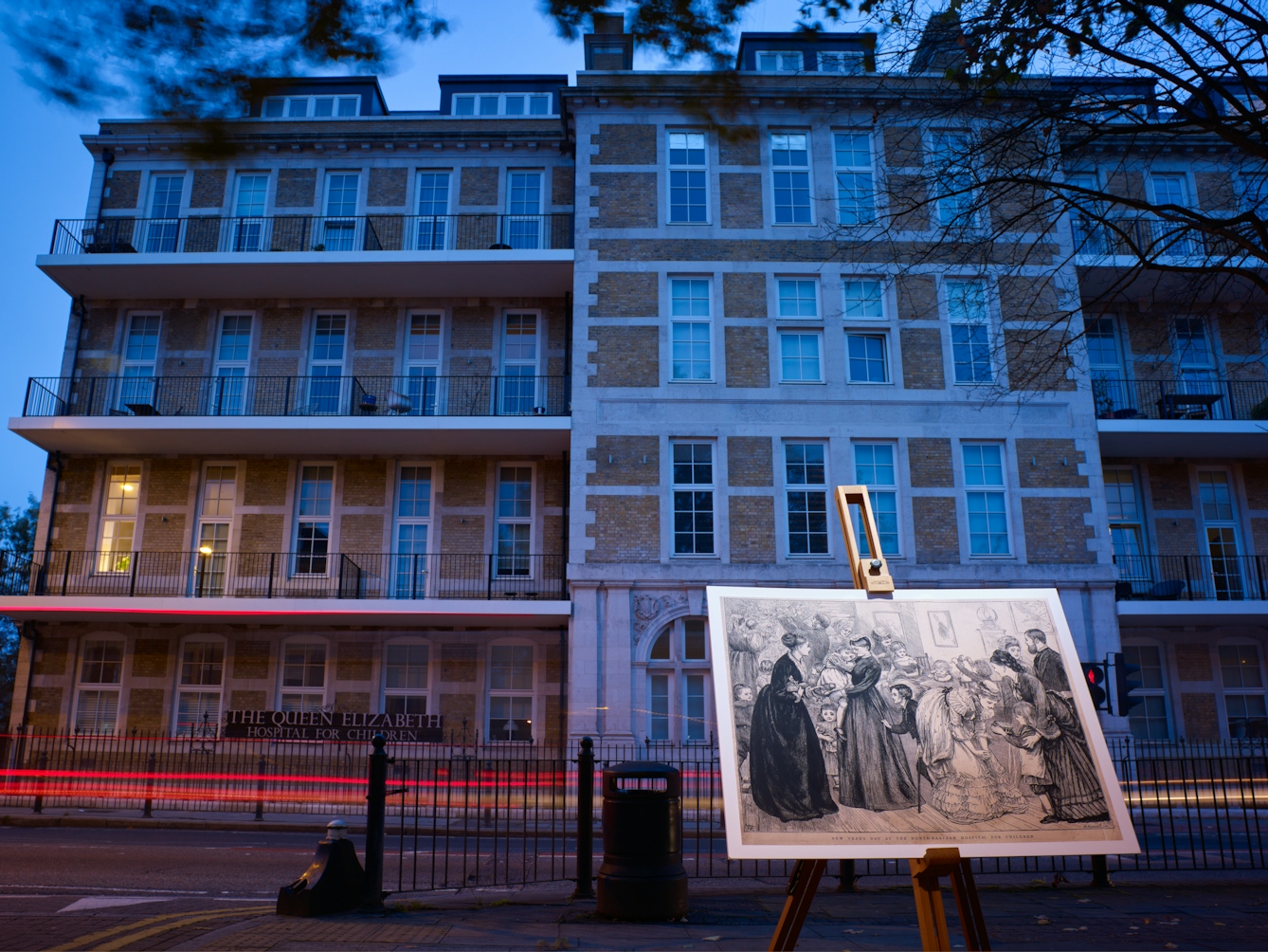 Photograph of the former Queen Elizabeth Children’s Hospital, Hackney Road with an historical image of the hospital displayed in front of it on a wooden artist's easel.