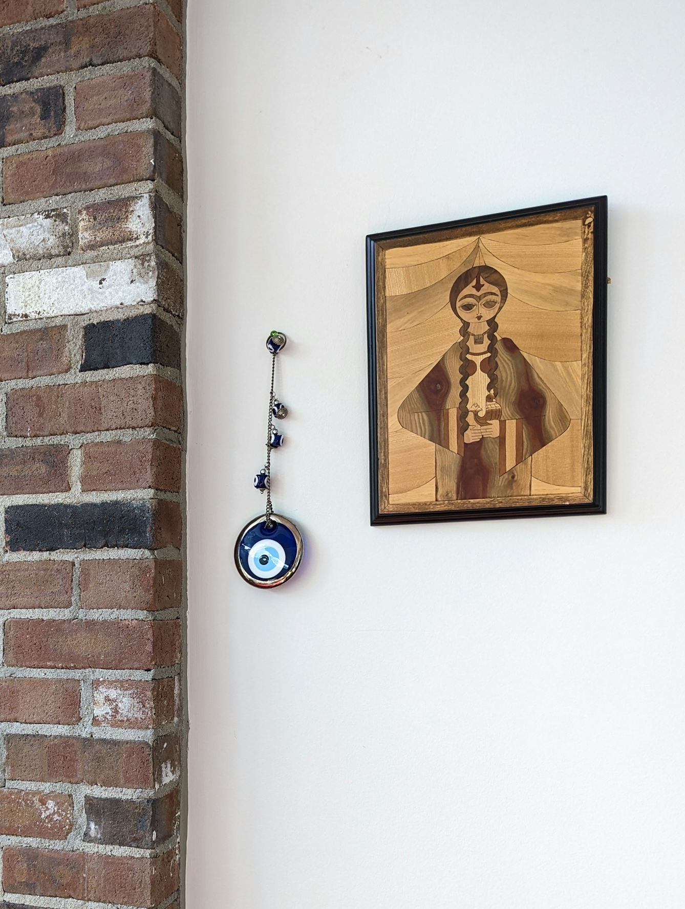 A large glass disc of concentric blue, white and black circles hangs on a single metal chain, with three smaller blue glass beads attached to the chain. The chain hangs on a plain wall between a marquetry artwork of a young wo,man and the edge of a brick pillar.