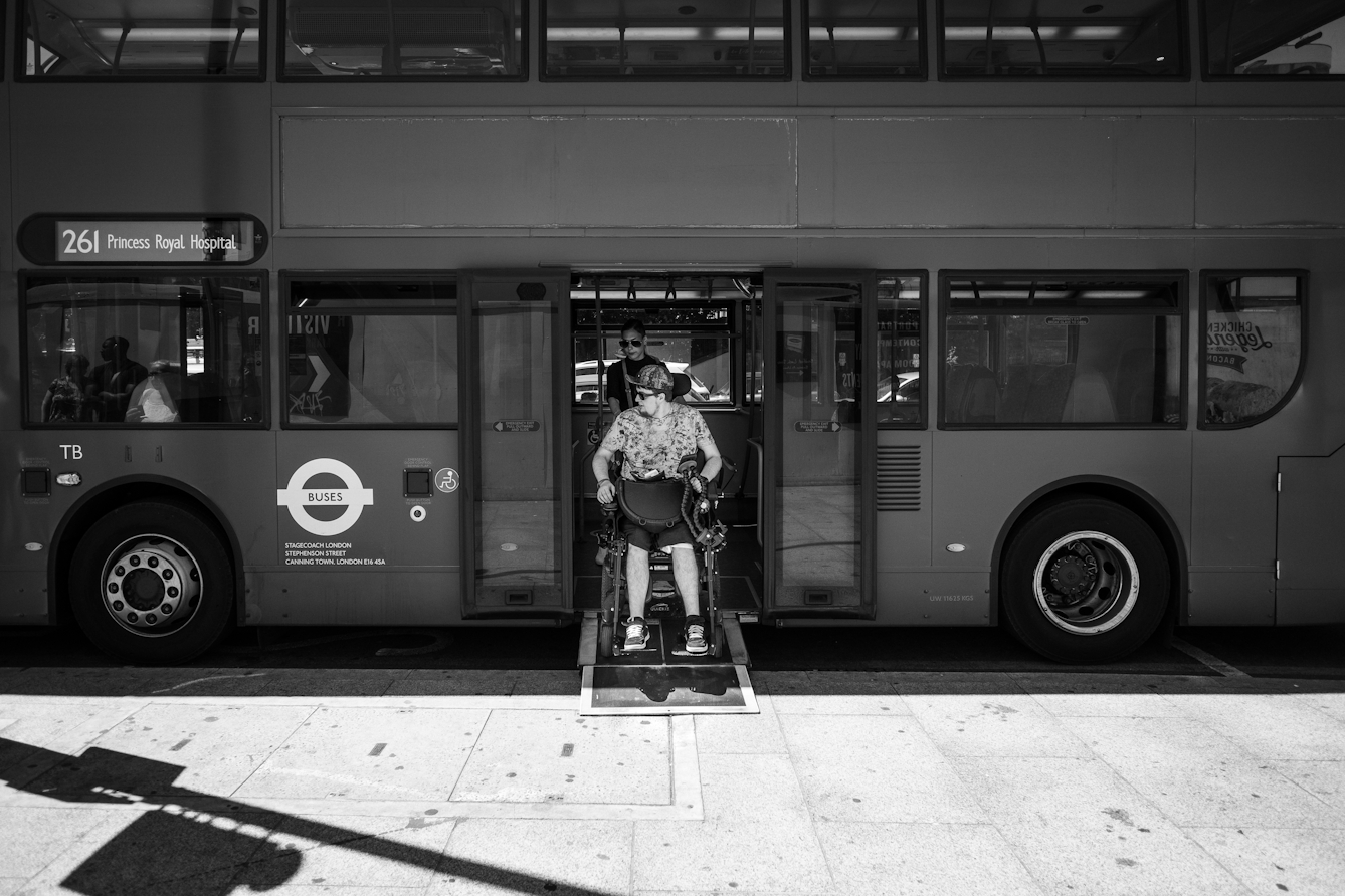 Black and white photograph of an individual reversing their wheelchair onto a London bus.