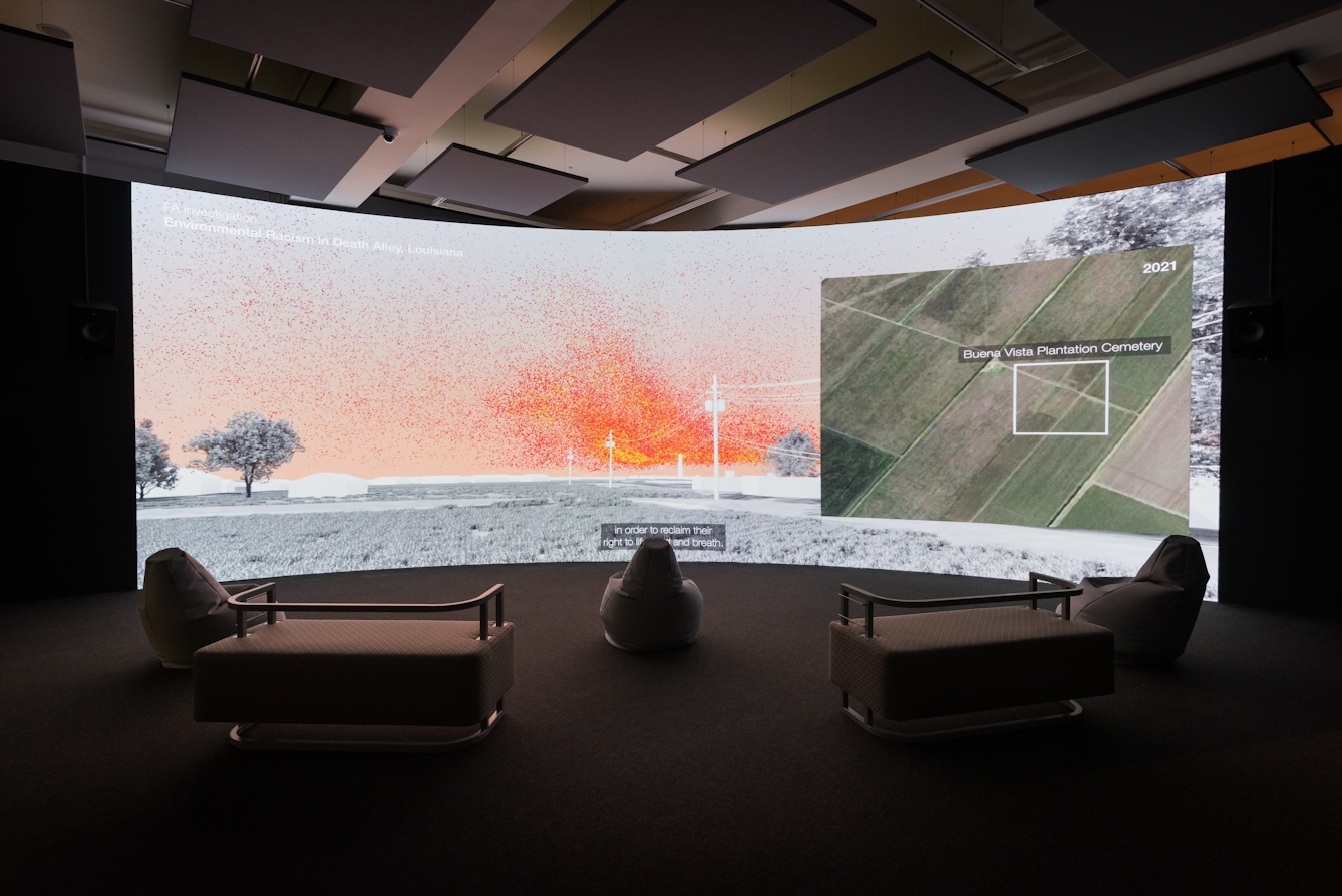 Photograph of a dark room with a very large curved screen and five empty chairs facing the screen. The screen shows a field with several trees and a telephone line running through it. To the right of the screen is an arial map of a number of green, partitioned plots of land. One section of land has an white square graphic surrounding it, and is labelled 'Buena Vista Plantation Cemetary'. 