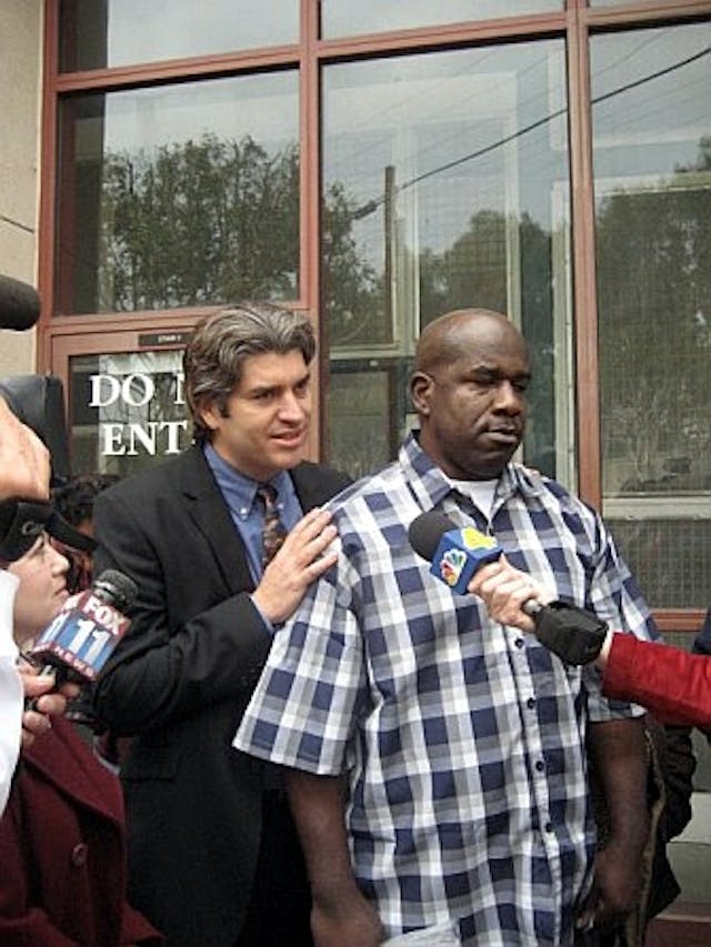 Image of a man with his hands on the shoulders of another man outside a courthouse. An arm holding a microphone points at them.