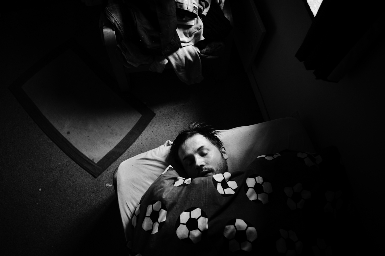 Black and white photograph showing a man lying on a mattress on the floor, his head appearing from a duvet with a football print on it.