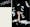 Photographic diptych, one large vertical image on the right and one smaller horizontal image on the left. The one on the right shows an arm and hand emerging vertically through a black velvet background, the fingers lightly curled. Around the arm, collected on the flat surface and floating down in the air are white feathers. The image on the left shows the word 'HARD' spelt out with white feathers, resting on a black background.