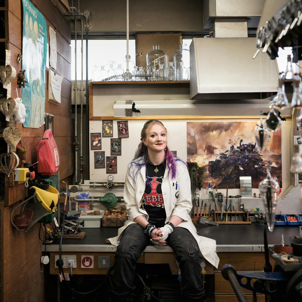 Picture of a woman sitting on a bench in a workshop wearing a white coat, with bright green sunglasses atop her head, blue and purple hair, and a black and red rock-logo t-shirt. Behind her are various glassblowing implements.