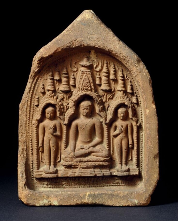 Photograph of a clay votive tablet. The round-headed design is impressed within a pointed-arched slab of terracotta. The figure of the Buddha is depicted seated beneath the tower of the Mahabodhi temple at Bodh Gaya, in eastern India. He is in padmasana pose in the Bhumisparsha mudra, flanked by standing figures of the Dipankara Buddha on the left and of Maitreya Buddha on the right. Each figure is placed within a niche. From the central niche also issue the branches of the Bodhi-tree. In the upper part of the tablet are a number of small votive stupas, and at the bottom is the "Ye dharma hetuprabhava..." or Buddhist prayer in Sanskrit characters. 
