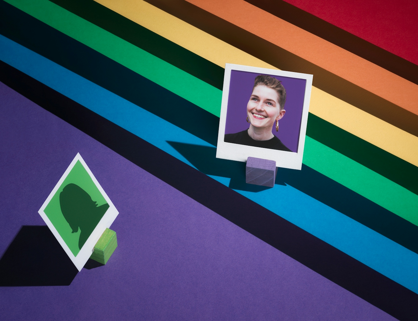 Photograph of a tiered set made up of the LGBTQ+ colours of red, orange, yellow, green, blue and purple.  The tiers form a diagonal line.  On the first tier, the blue one, the portrait of a woman against a purple background is framed in a small white frame.  Below this image, to the left, an anonymous figure on a green background is facing towards her.