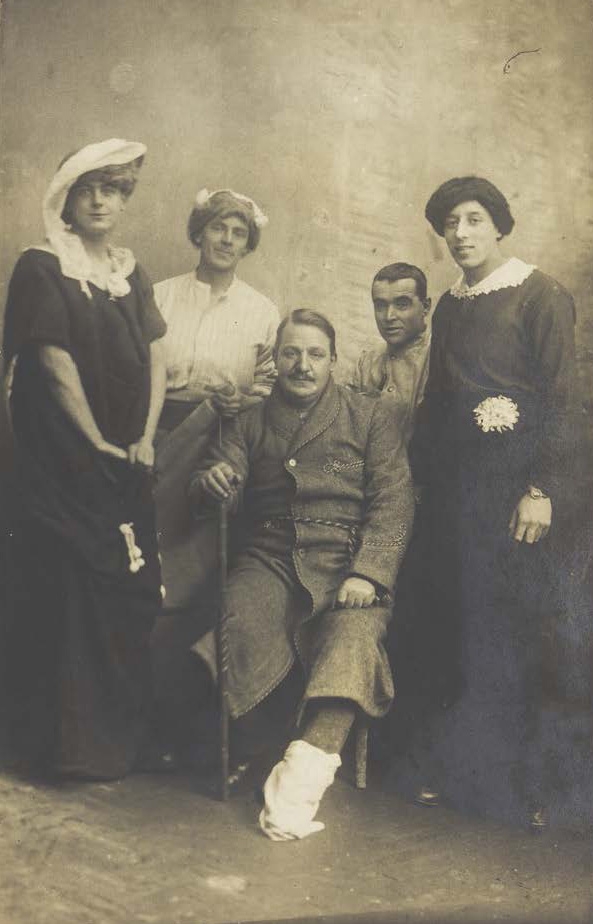 Four soldiers posing, three in drag, with an injured soldier seated in the centre, photographic postcard, c.1910-19