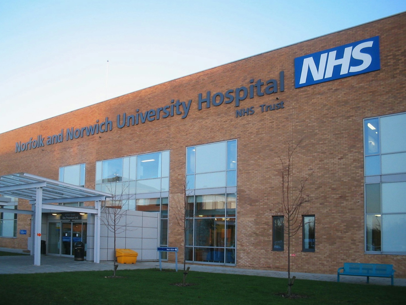 Colour photograph of the entrance to the Arthur South Day Procedure Unit at the Norfolk and Norwich University Hospital. Building is made of brick with large glass windows and a glass entranceway, and has a sign across the top with the name of the hospital trust and with NHS logo.