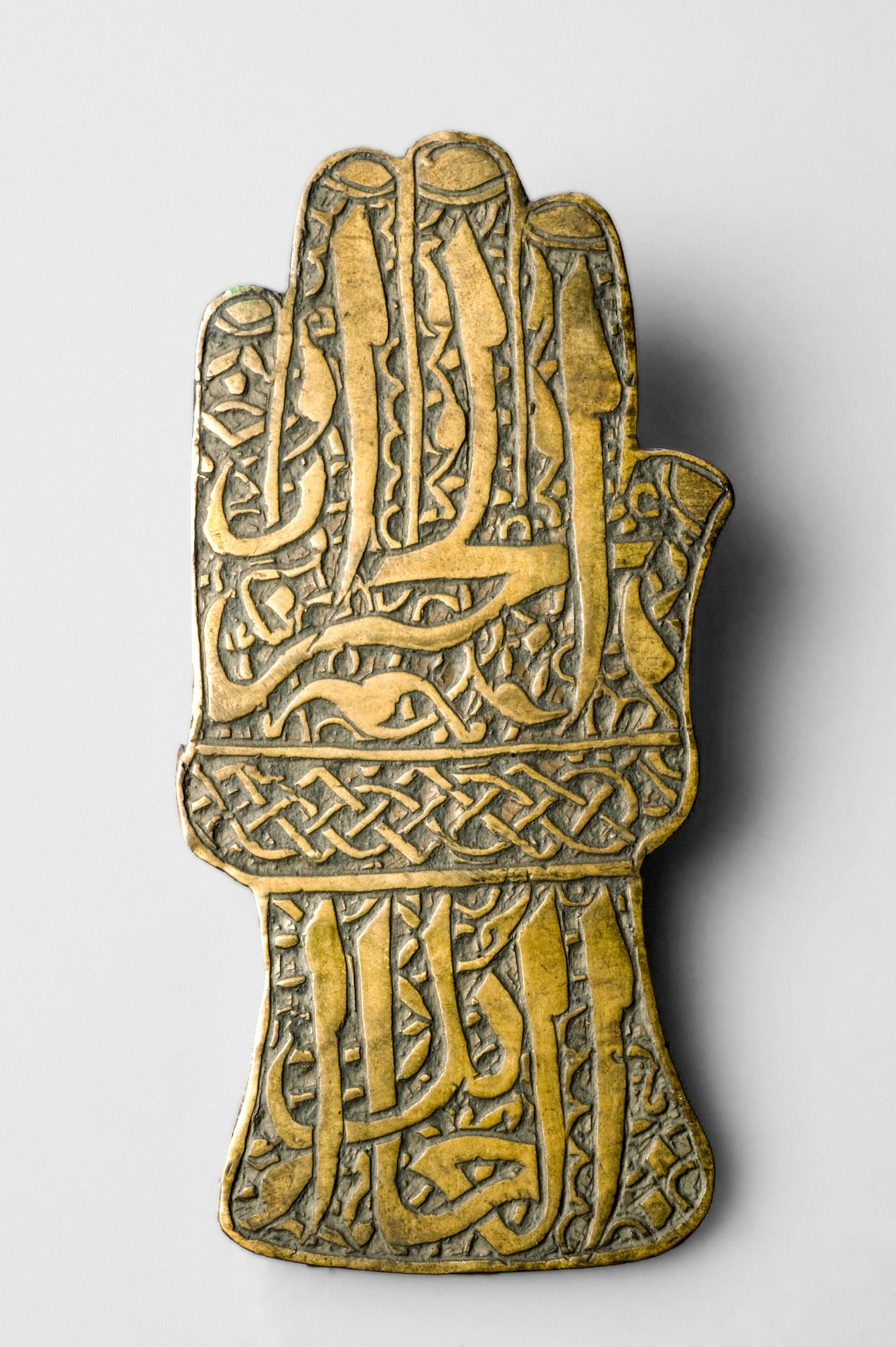 A brass amulet in the shape of the a the left hand, palm facing down. The surface is ornately inscribed with details fo the fingers, thumb and fingernails and arabic text, interspersed with decorative patterns.