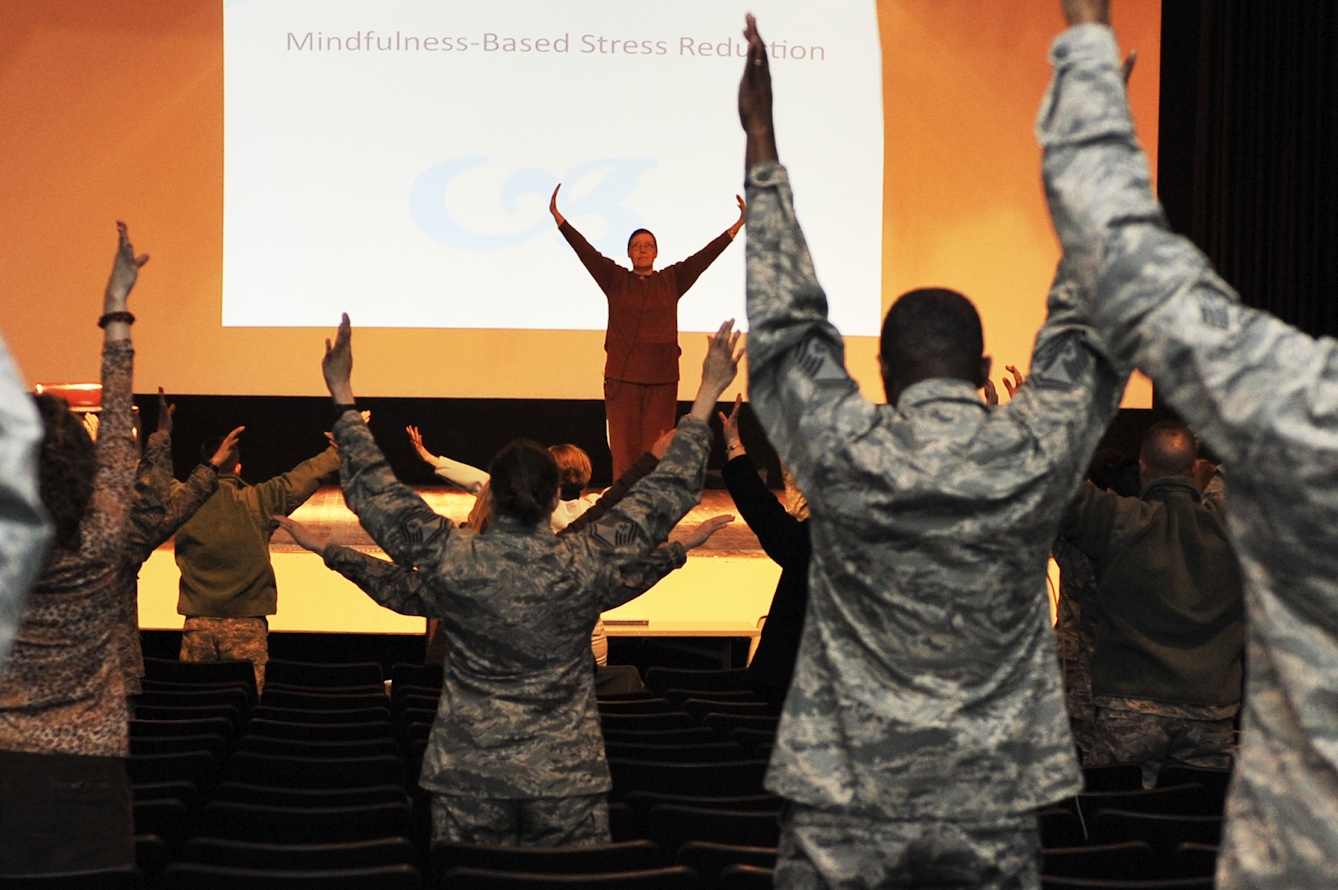 In front of a screen reading "Mindfulness-Based Stress Reduction", a woman with her arms raised leads a class of people in military fatigues copying her movements.