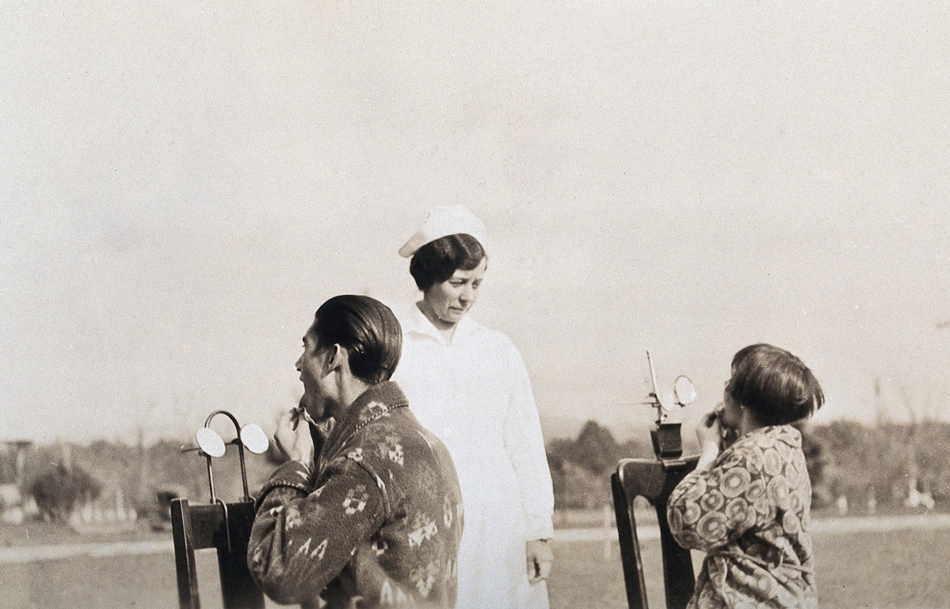 Black and white photograph of a nurse in uniform observing tow patients wearing dressing gowns as they carry out their helio treatment in the middle of an empty field. A man and a girl aged around 12-14 are sat on chairs with their backs to the camera but infront of the nurse. The chairs have two small round mirrors mounted on them, reflecting light into thier mouths. The patients have their mouths wide open and hold thier tongues out with their hands. 