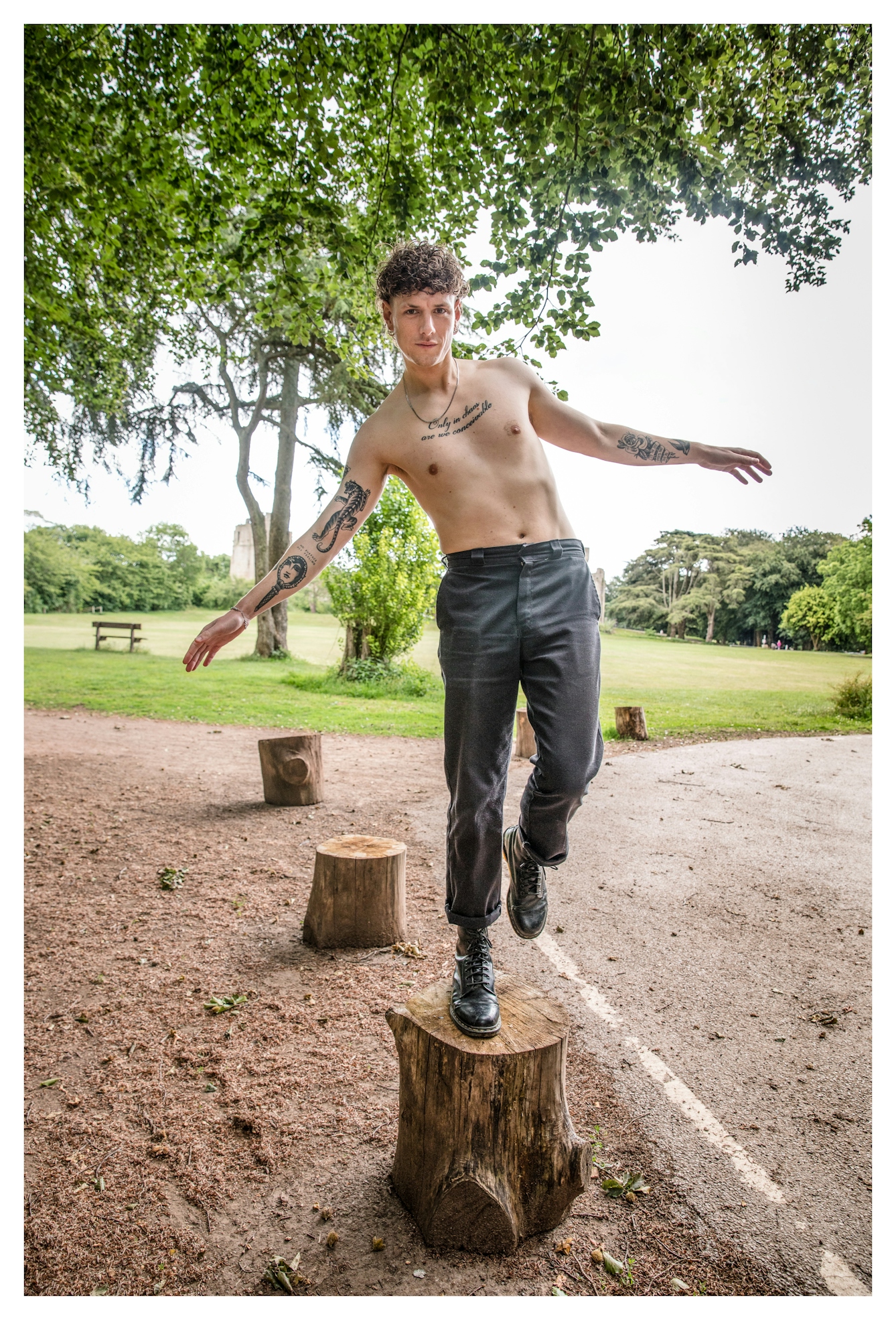 Photograph of a man balancing on a cut tree stump in open parkland. He has his shirt off, reaving his upper torso which is adorned with several tattoos. 
His arms are outstretched, one leg raised off the ground and he's looking straight to camera. Behind him is a large expanse of grass, trees, paths and benches.