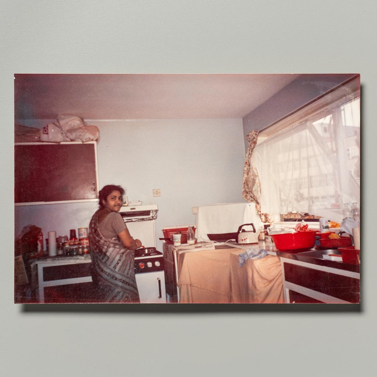 Photograph of a family photographic print resting on a grey background. The photo shows a mother in a kitchen tending to a pan on the stove. She is looking over her shoulder into the camera.