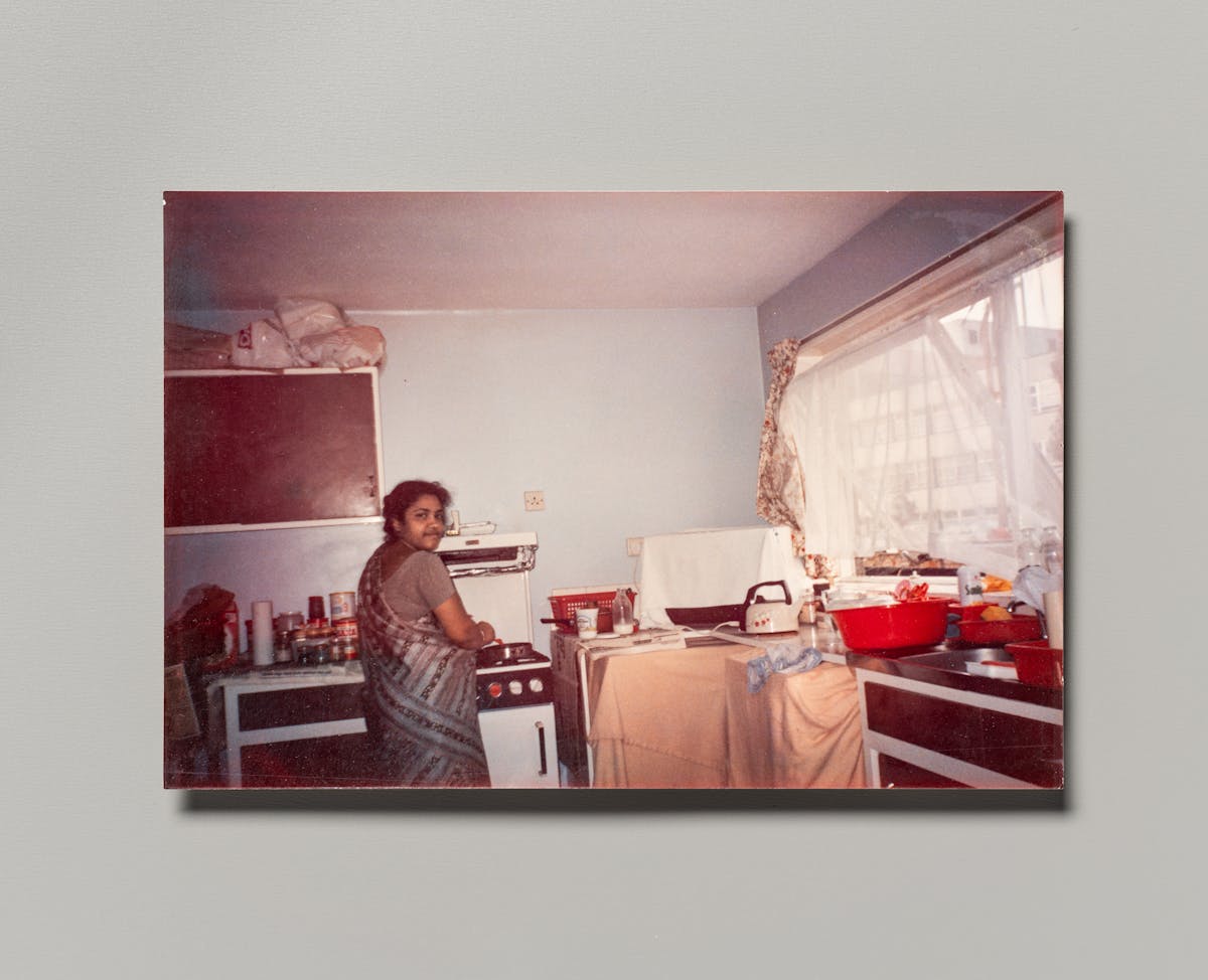 Photograph of a family photographic print resting on a grey background. The photo shows a mother in a kitchen tending to a pan on the stove. She is looking over her shoulder into the camera.