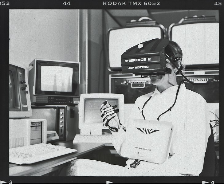 Black and white photograph of a person wearing a large headset labelled Cyberface 2 LEEP Boston, with wires linking to a large rectangular device on their chest and a glove on their right hand. In the background are four chunky 1980s computer monitors and two keyboards.
