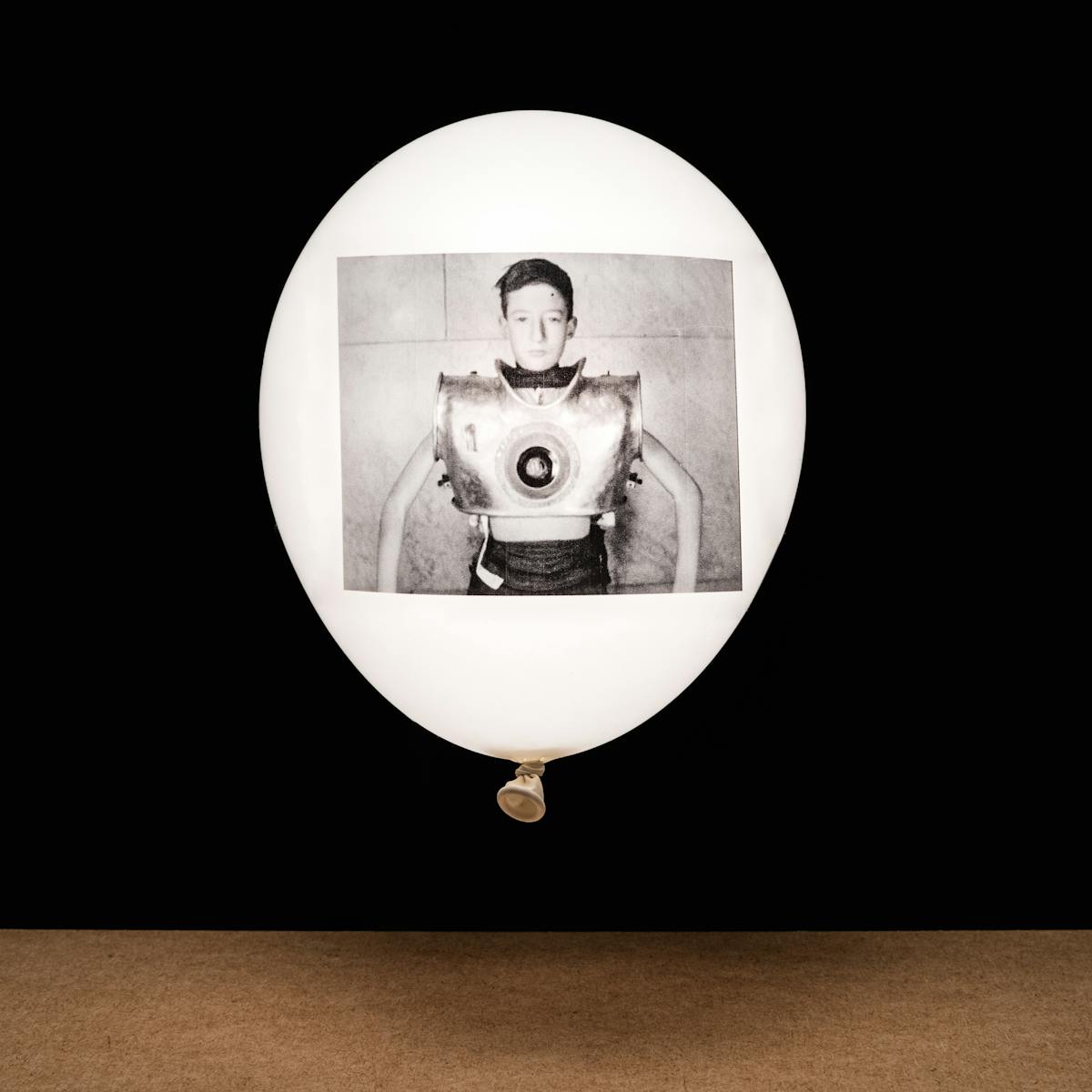 Photograph of a white inflated balloon against a black background, floating vertically above a wooden tabletop horizon line. The balloon looks like it is illuminated from within. On the side of the balloon is a rectangular, monochrome archive film still. The still shows a young boy facing the camera. His chest in encased in large metal contraption which is artificial ventilator.